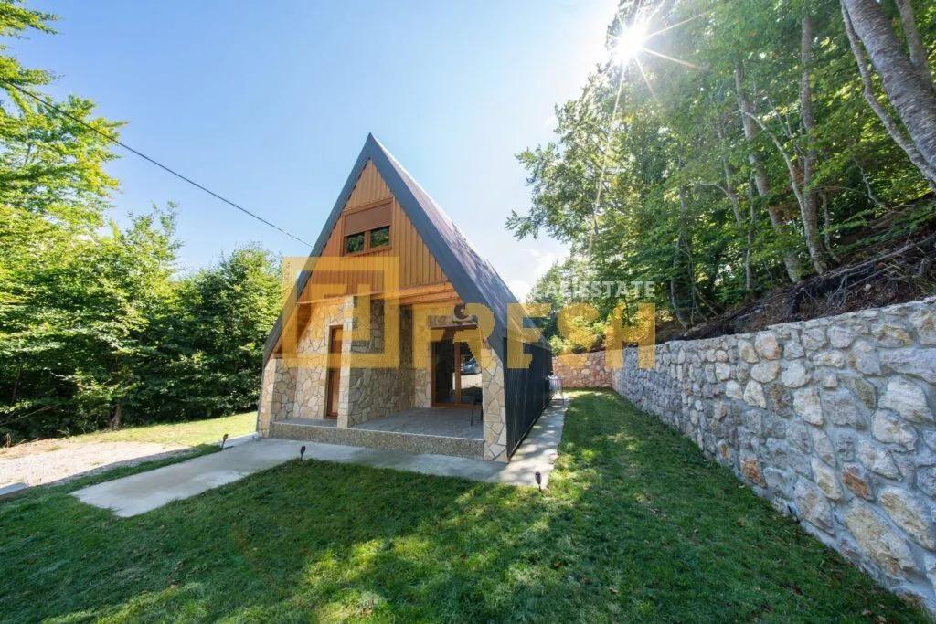 Pogled na drvenu vikendicu okruženu šumom i kamenim zidom.
