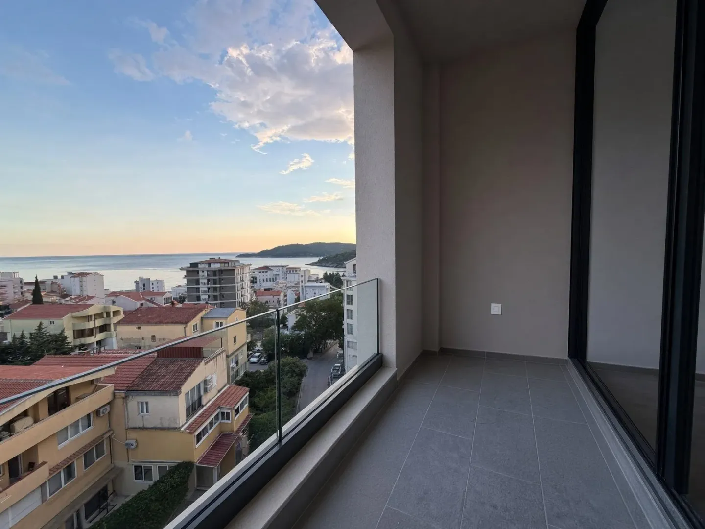 Nenamješten balkon sa staklenom ogradom i pogledom na grad i more.