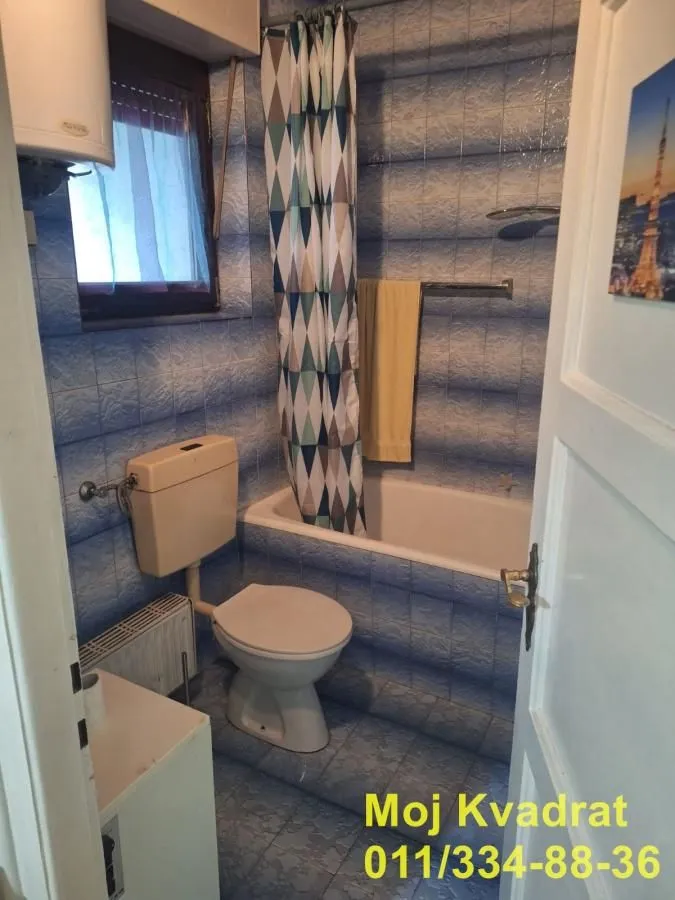 Manje kupatilo sa kadom, wc šoljom i prozorom obloženo plavim pločicama.