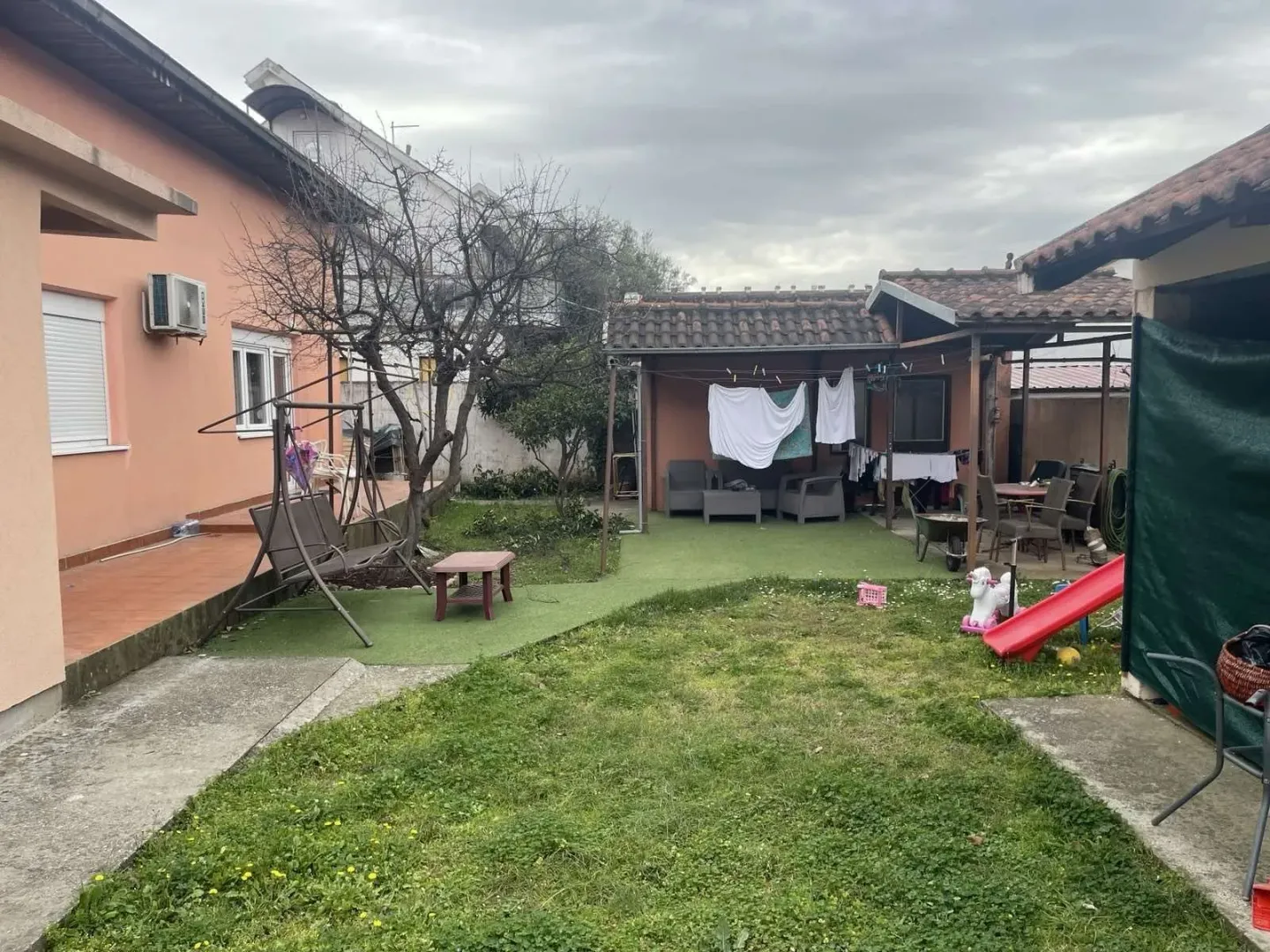 Dvorište porodične kuće sa travnjakom, baštenskim namještajem i pomoćnim objektom.