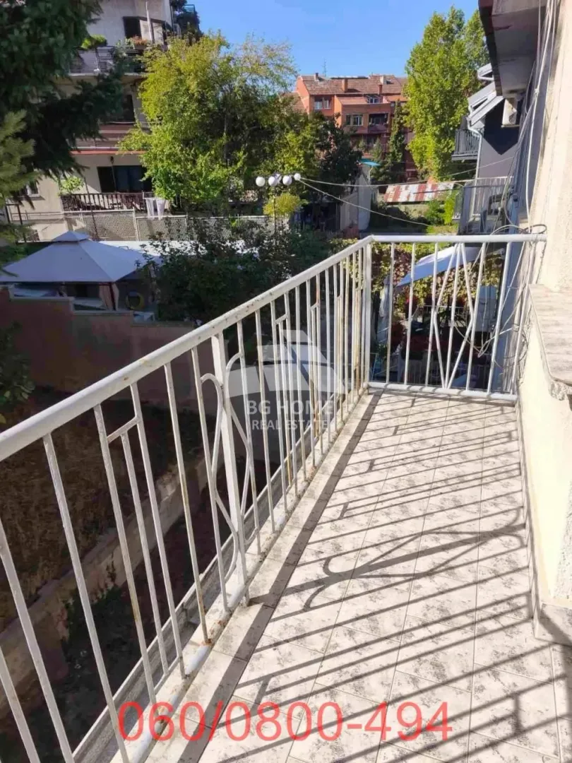 Pogled sa popločanog balkona sa belom ogradom na okolne zgrade i zelenilo.
