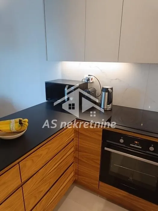 Manja kuhinja sa drvenim elementima, crnom radnom površinom i ugrađenim kućnim aparatima.