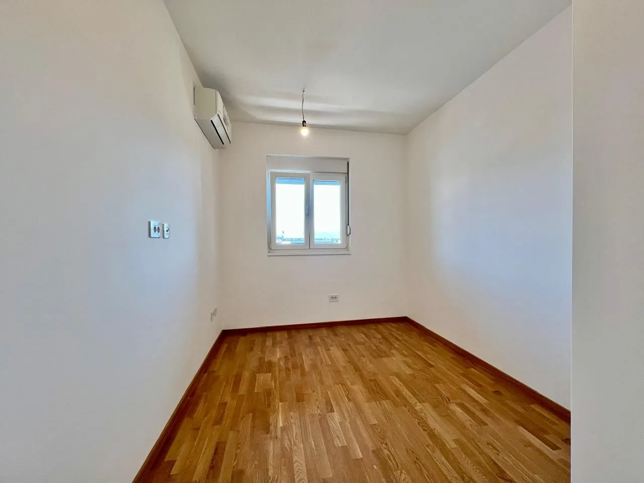 Prazna soba sa bijelim zidovima, parketom i prozorom.