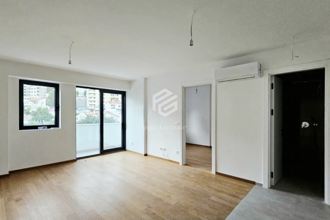 Prazna soba sa drvenim podom, bijelim zidovima i izlazom na balkon.