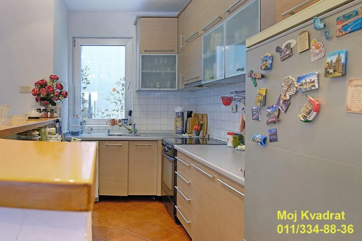 Manja kuhinja sa drvenim elementima, sudoperom ispod prozora i frižiderom sa magnetima.