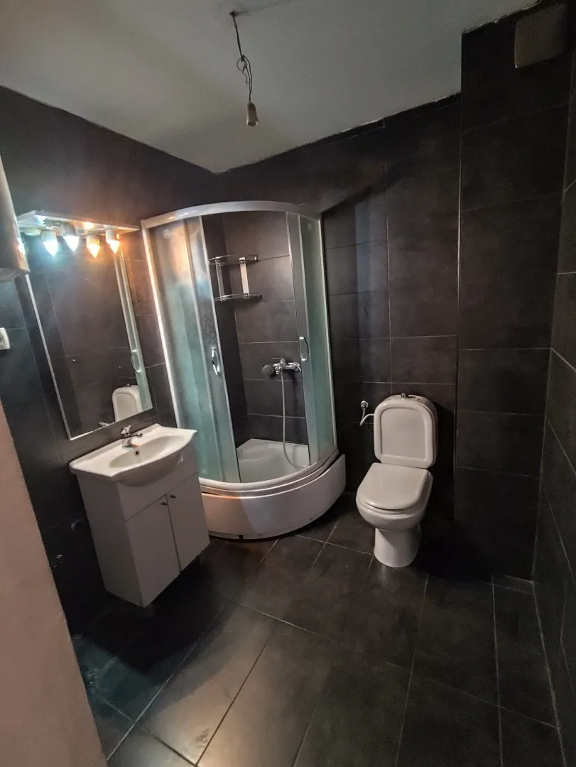 Kupatilo sa tamnim pločicama, tuš kabinom, umivaonikom i wc šoljom.