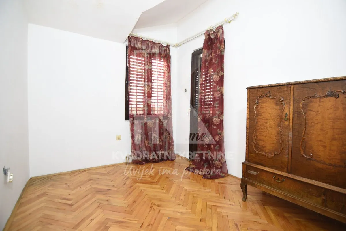 Empty room with hardwood parquet flooring, an antique wooden cabinet, and windows with red curtains.