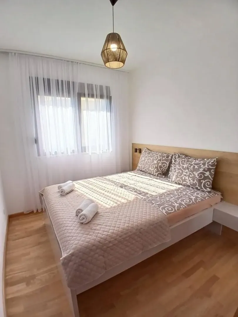 Uređena spavaća soba sa bračnim krevetom, drvenim podom i prozorom sa bijelim zavjesama.