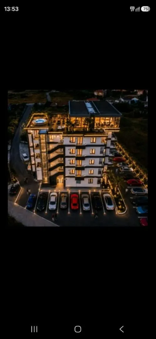Noćni pogled iz vazduha na modernu zgradu sa osvetljenim parkingom i krovnom terasom.