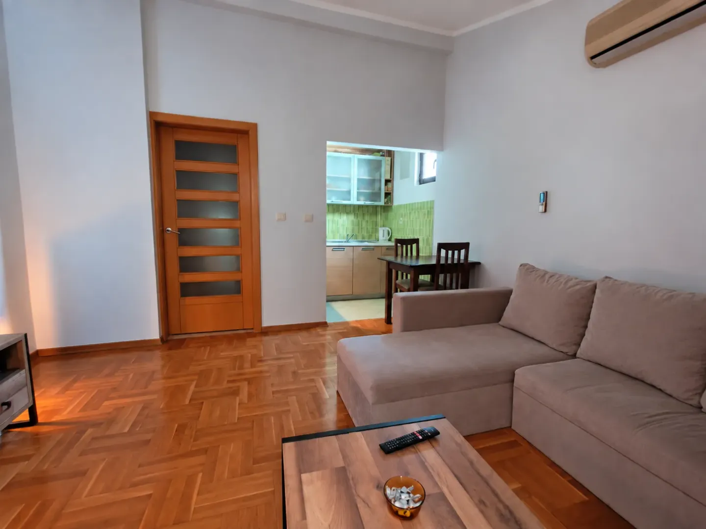 Living room with a sectional sofa, wooden parquet flooring, and a view of the kitchen.