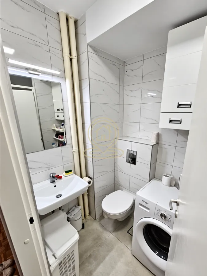 Manje kupatilo sa belim pločicama, umivaonikom, WC šoljom i veš mašinom.