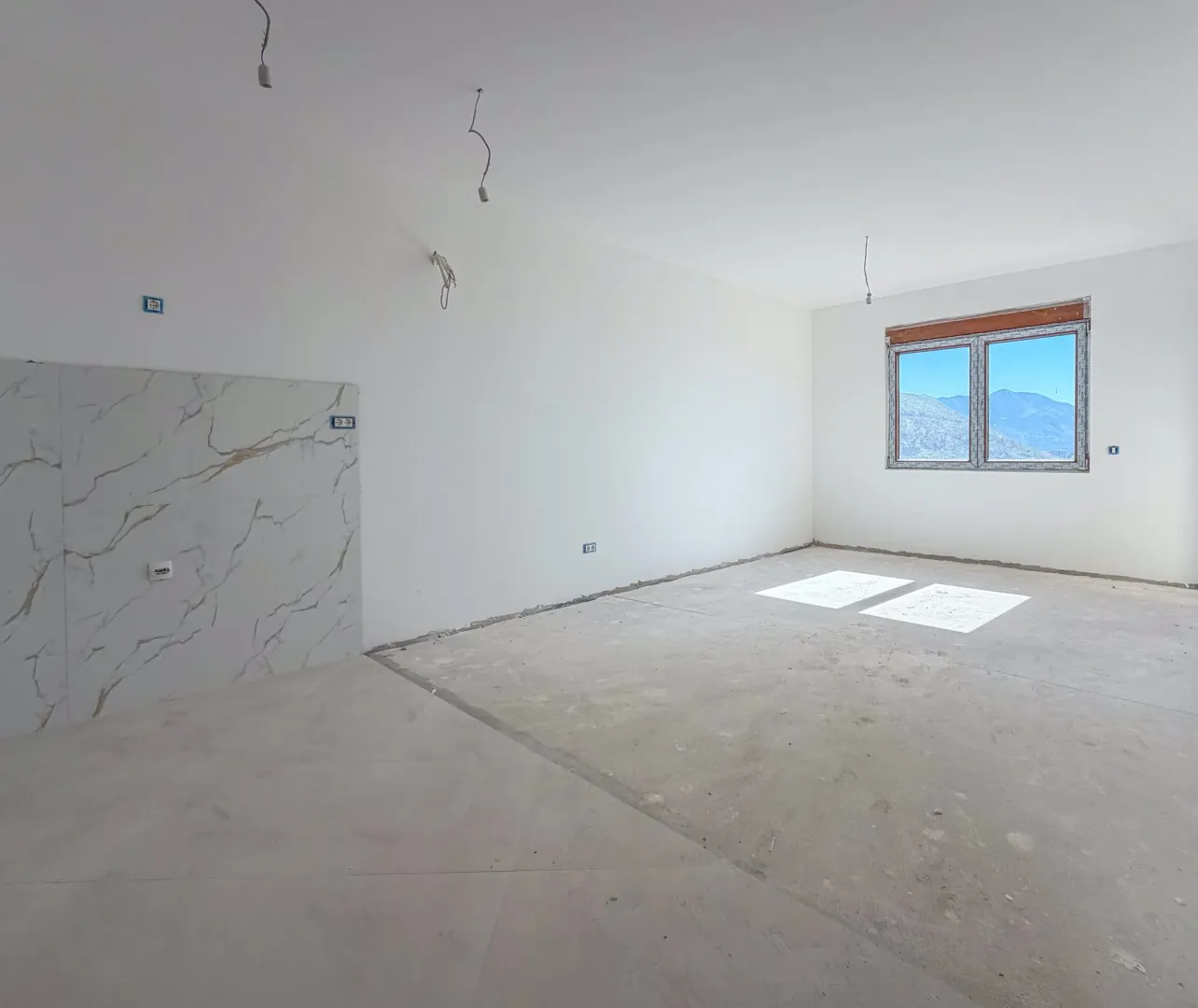 Nezavršena prostorija sa bijelim zidovima, pločicama na dijelu zida i prozorom koji gleda na planine.
