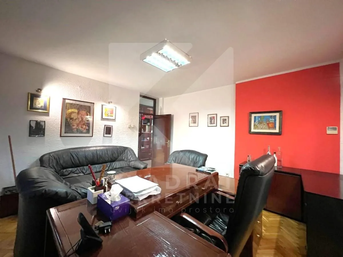 Kancelarija sa drvenim radnim stolom, crnim kožnim foteljama i umjetničkim slikama na zidu.