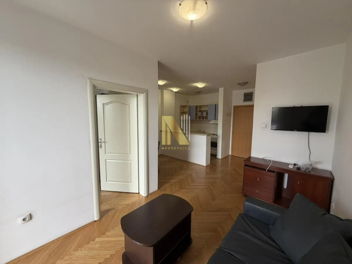 View of the living room with a sofa and TV, with a passageway leading to the kitchen and the entrance door.