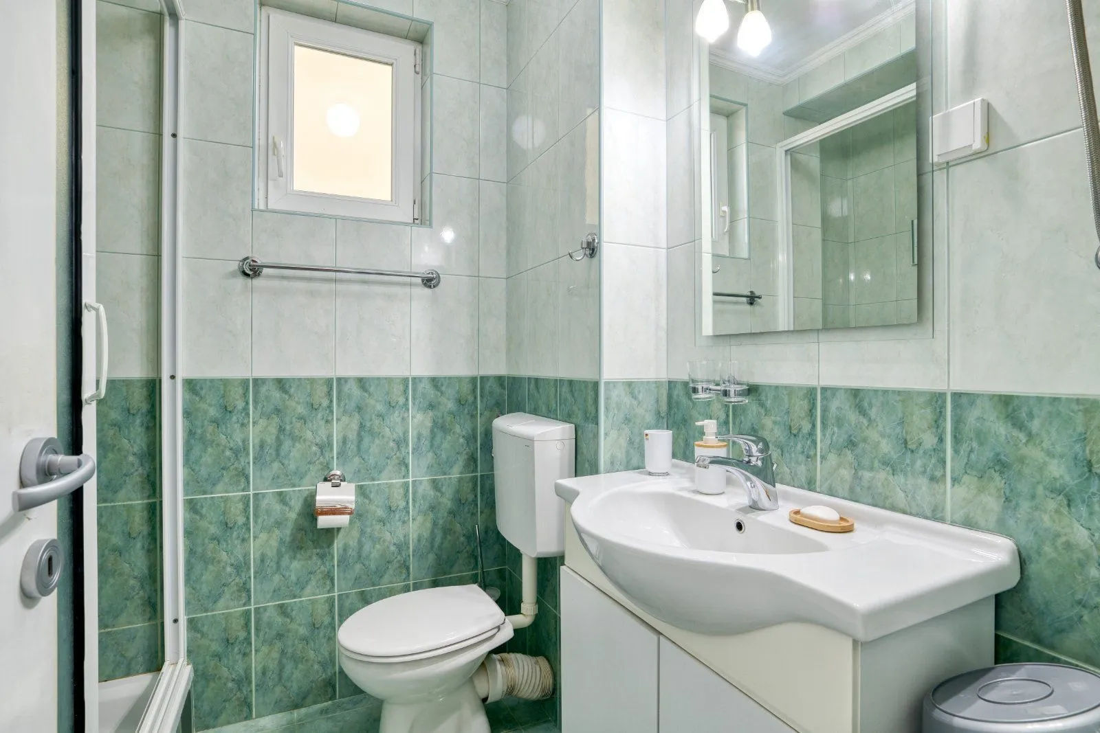 Compact bathroom tiled in green with a washbasin, toilet, and shower enclosure.