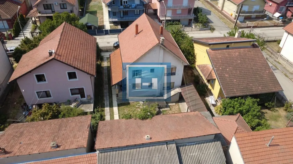 Pogled iz vazduha na kuću i okolno naselje sa krovovima i dvorištima.