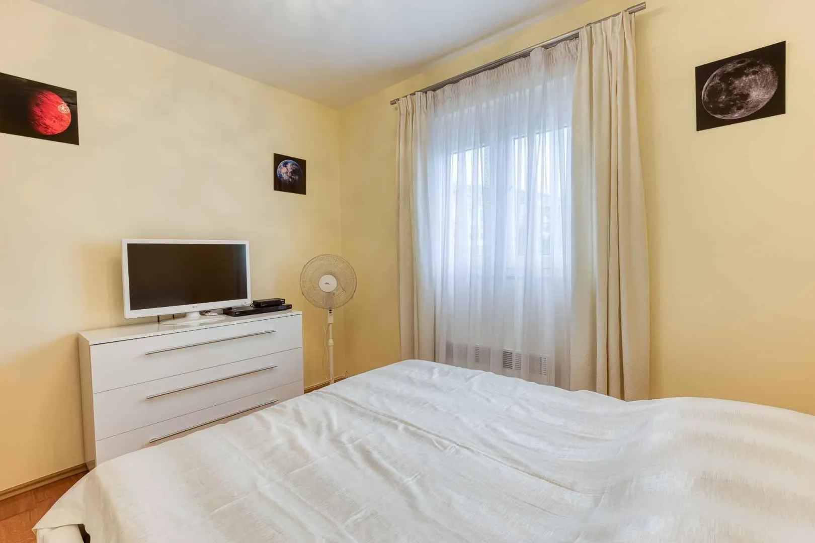 Furnished bedroom with a double bed, dresser, television, and a window with white curtains.