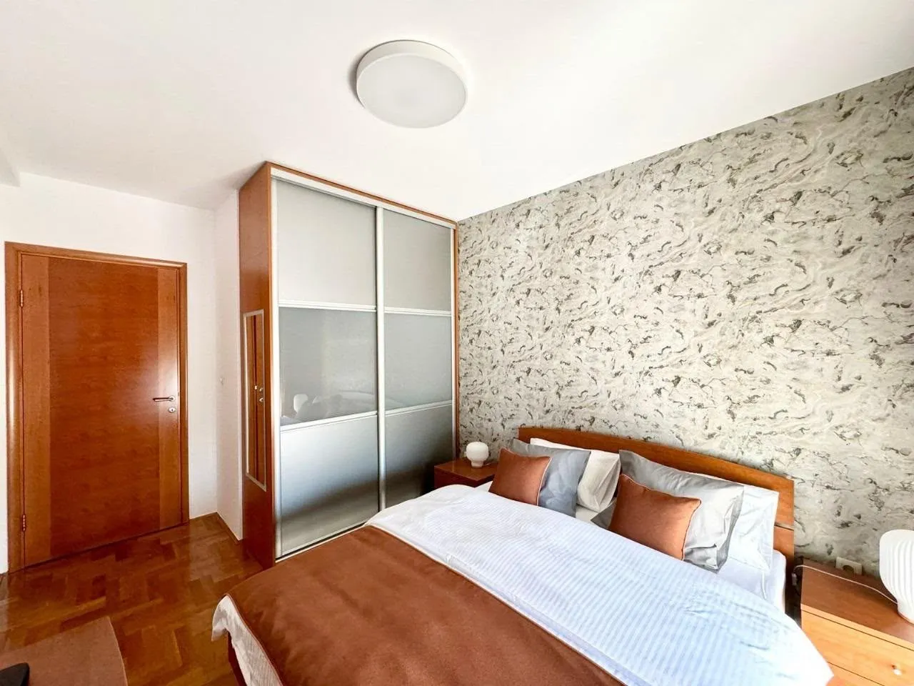 Furnished bedroom with a double bed, wardrobe, and patterned wall covering.