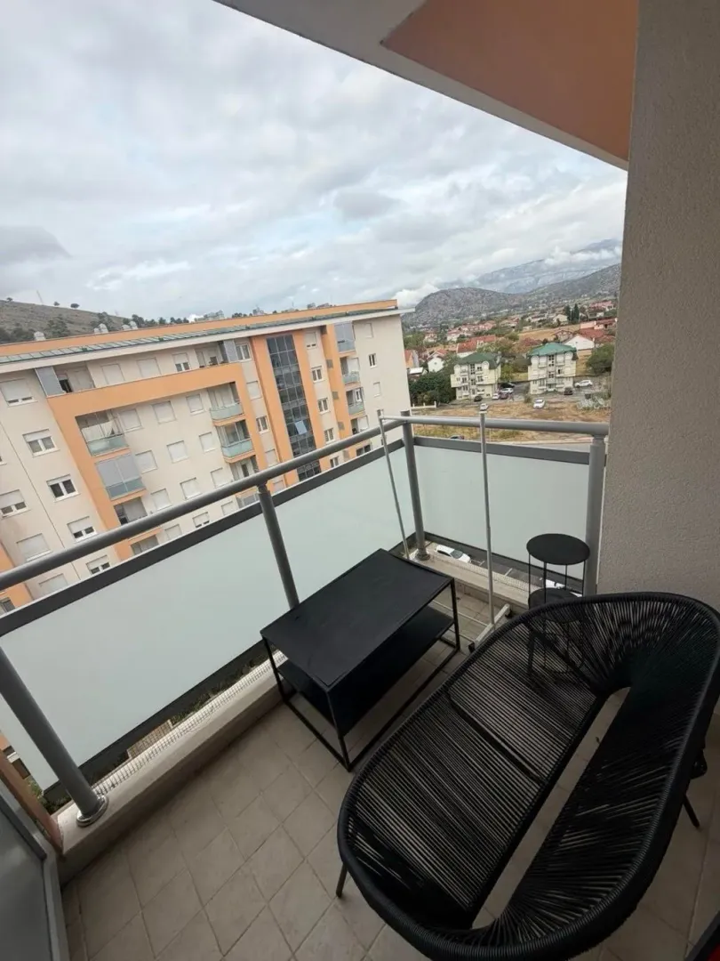 Nenamešten balkon sa crnim stolom i klupom, i pogledom na okolne zgrade i pejzaž.
