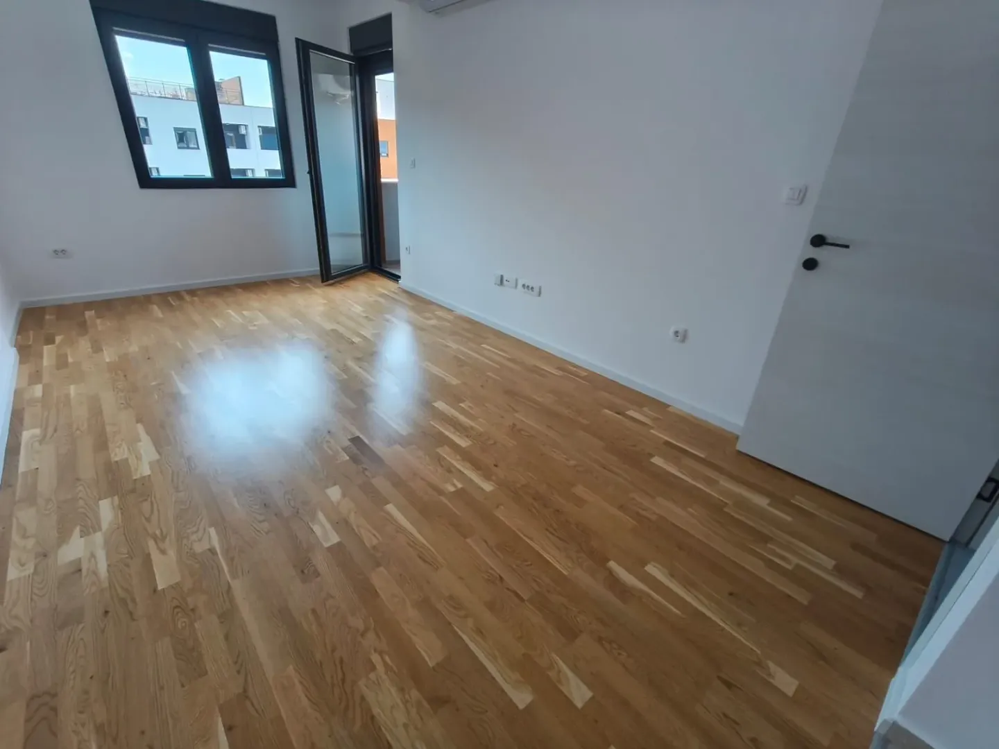 Prazna soba sa drvenim parketom, bijelim zidovima i izlazom na terasu.