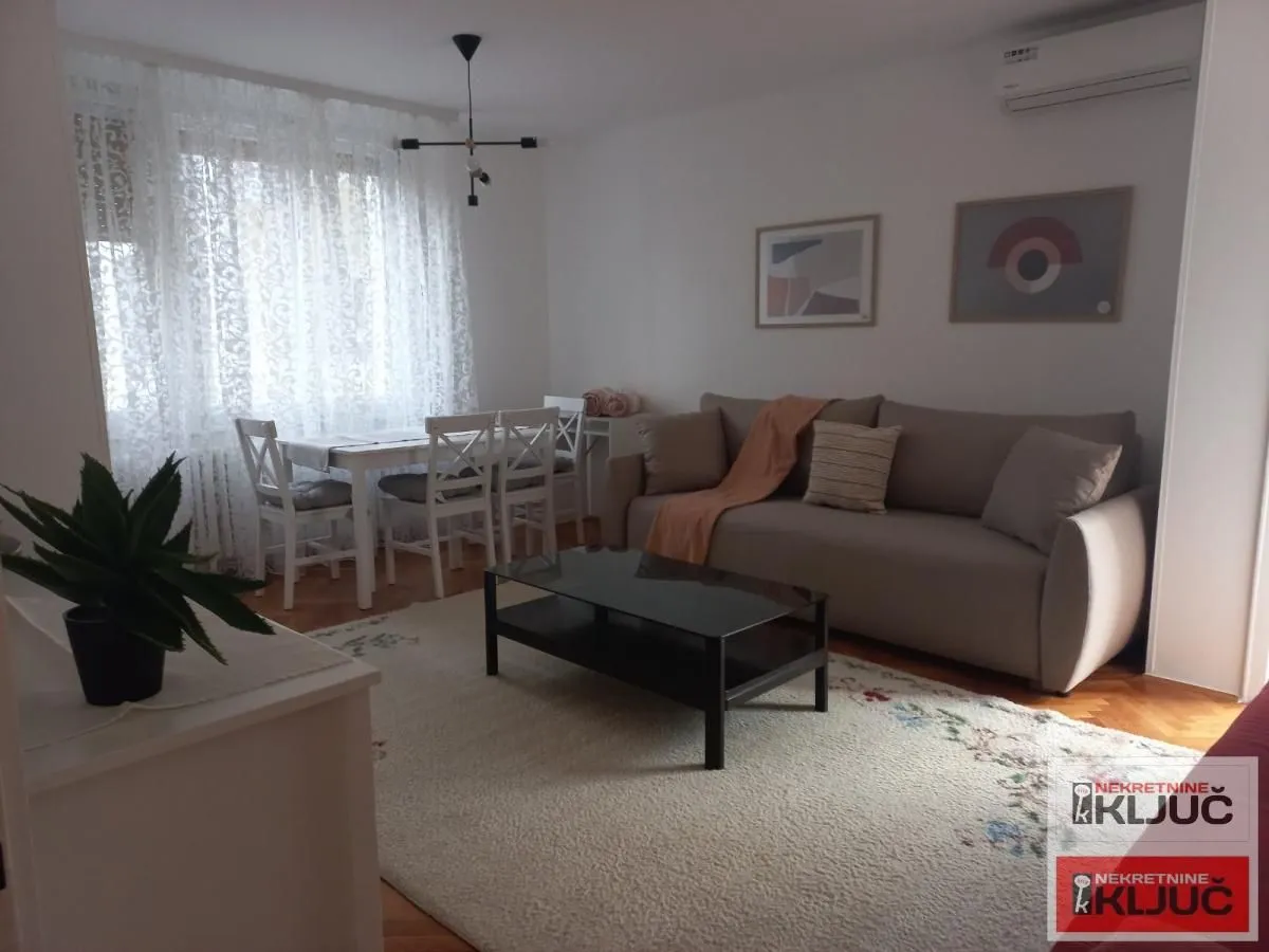 Dnevna soba sa garniturom, stočićem i trpezarijskim delom pored prozora.