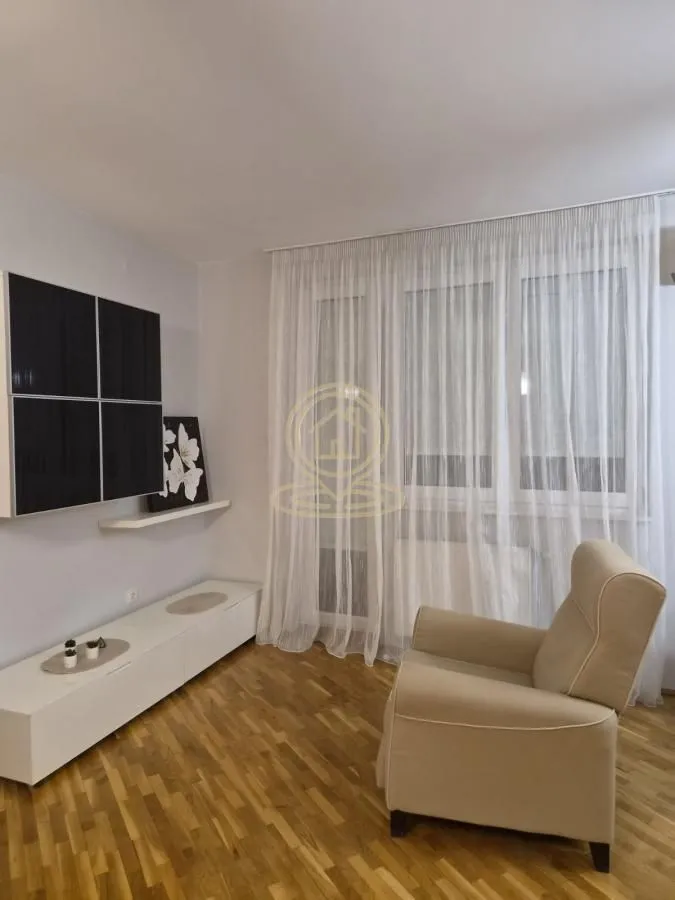 Dnevna soba sa foteljom, TV komodom i prozorima sa belim zavesama.
