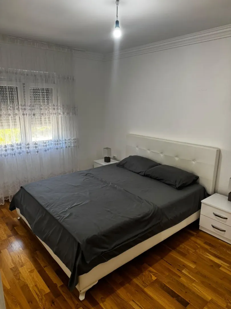 Spavaća soba sa bračnim krevetom, bijelim noćnim stočićima i prozorom sa zavjesama.