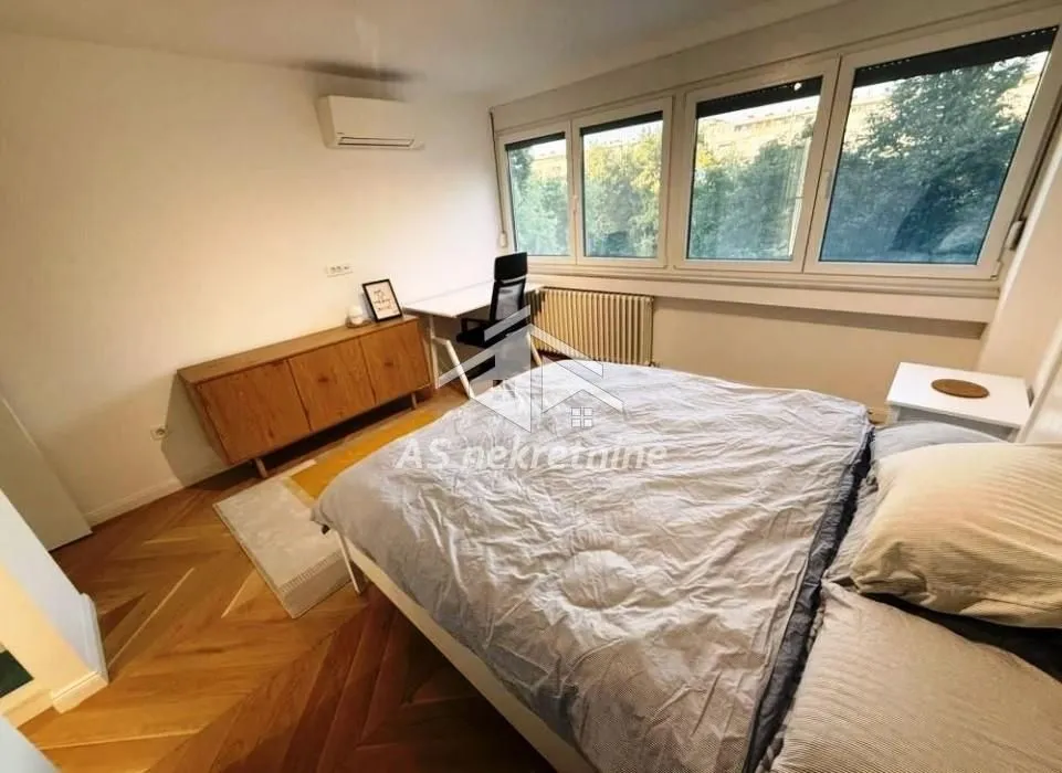 Nameštena spavaća soba sa bračnim krevetom, radnim stolom i velikim prozorima.