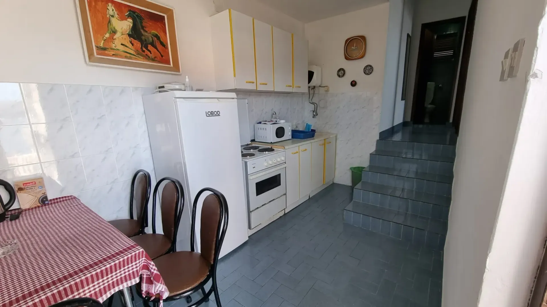 Manja kuhinja sa bijelim elementima, frižiderom i trpezarijskim stolom, pored stepeništa.