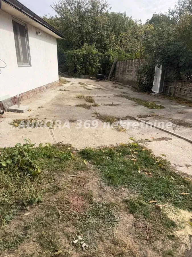 Betonsko dvorište pored kuće sa ogradom i zelenilom.