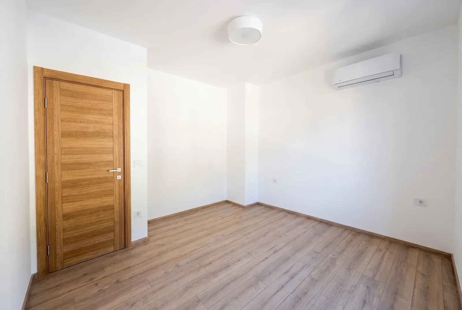 Empty room with white walls, wooden flooring, and an air conditioning unit.