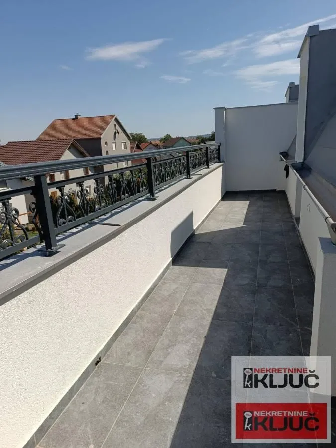 Prostrani popločani balkon sa crnom metalnom ogradom i pogledom na okolinu.