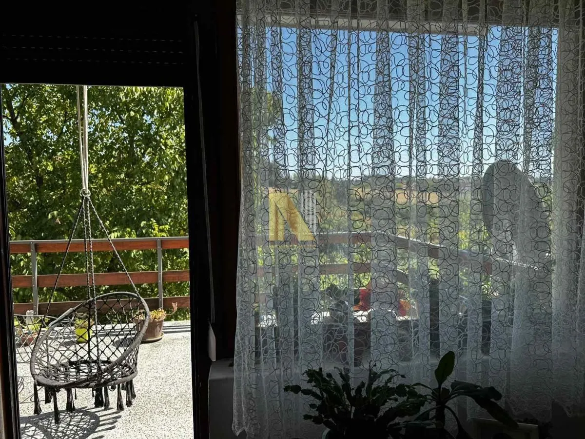 Pogled kroz prozor i vrata na terasu sa visećom stolicom i zelenilom.