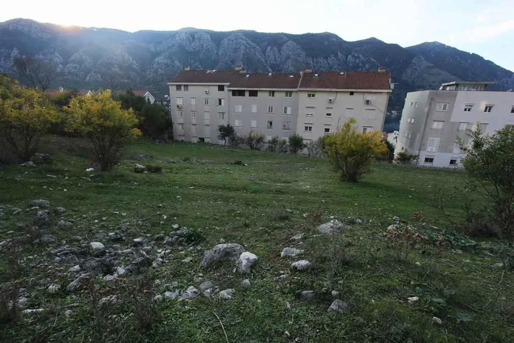 Travnati plac sa stambenim zgradama u pozadini i planinskim vijencem.