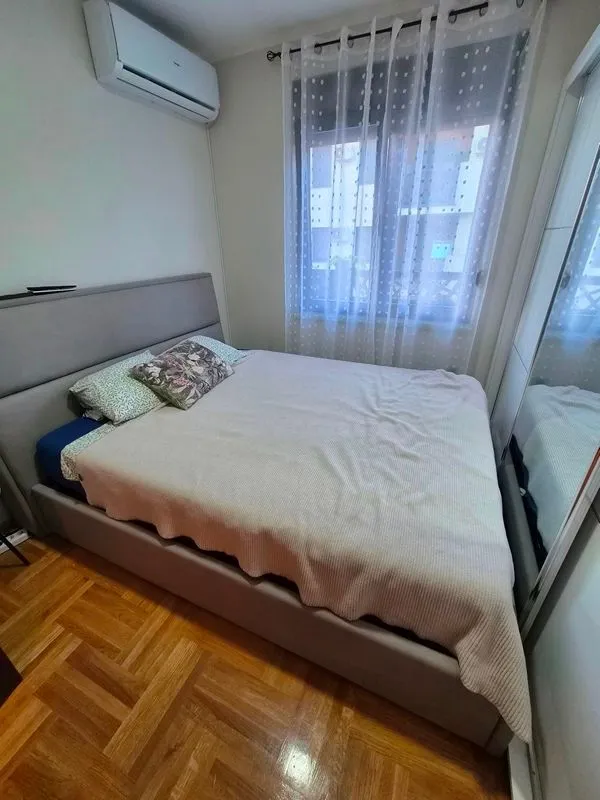 Nameštena spavaća soba sa bračnim krevetom, klima uređajem i prozorom sa zavesama.