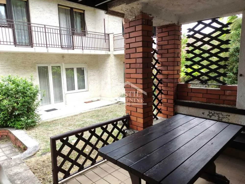 Natkrivena terasa sa drvenim stolom i pogledom na dvorište zgrade.