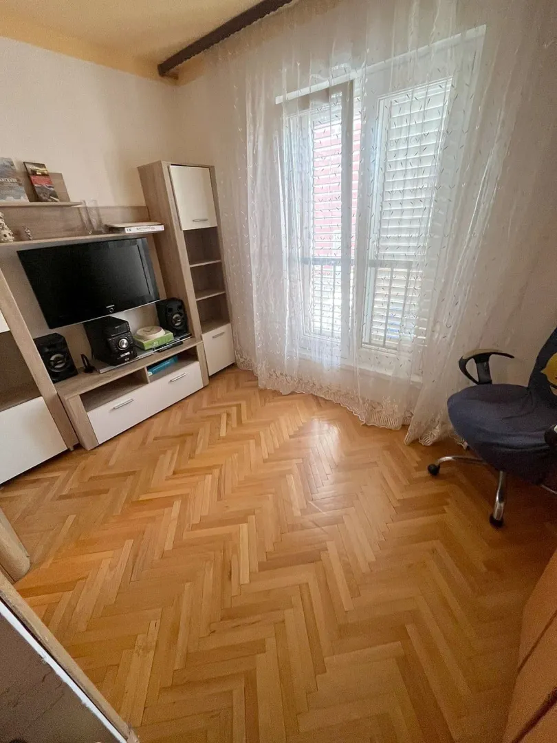 Dnevna soba sa drvenim podom, TV komodom i prozorom sa zavjesama.