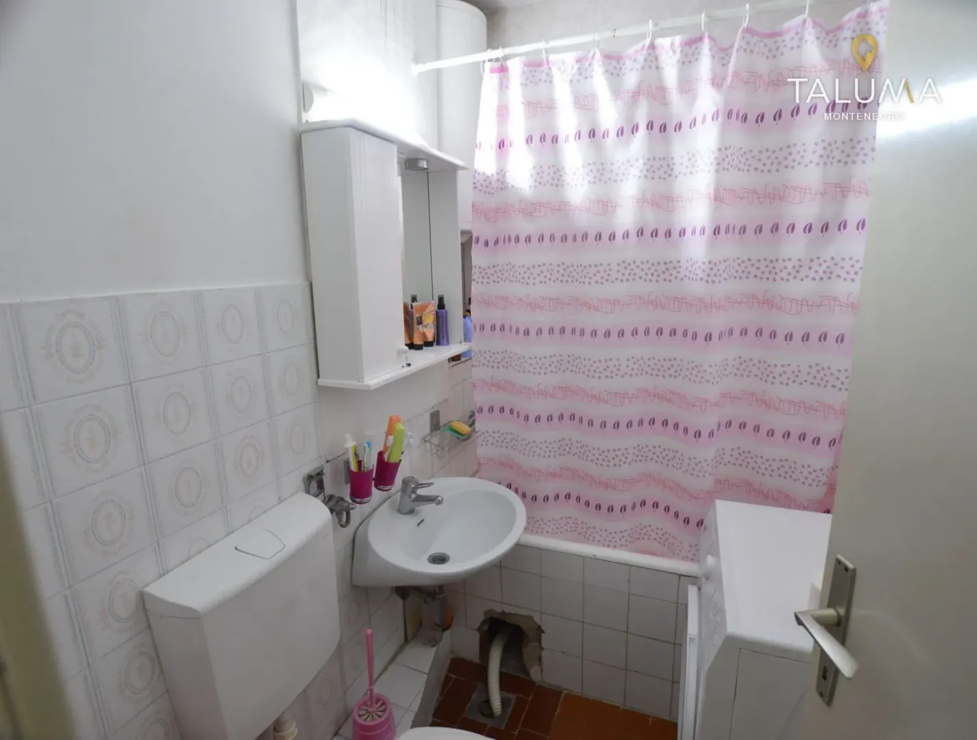 Manje kupatilo sa umivaonikom, WC šoljom, kadom sa zavesom i ormarićem sa ogledalom.