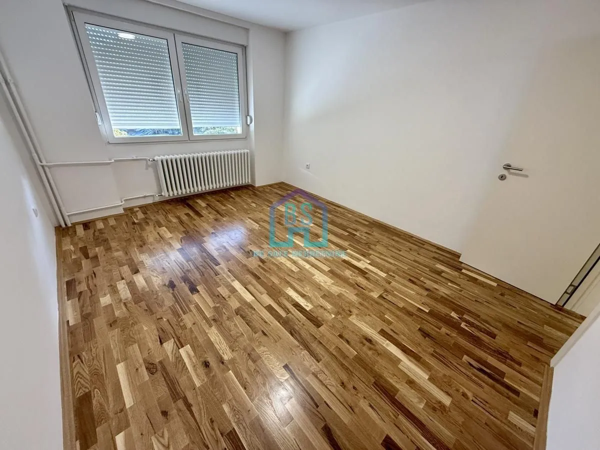 Prazna neopremljena soba sa drvenim parketom i prozorom sa roletnama.