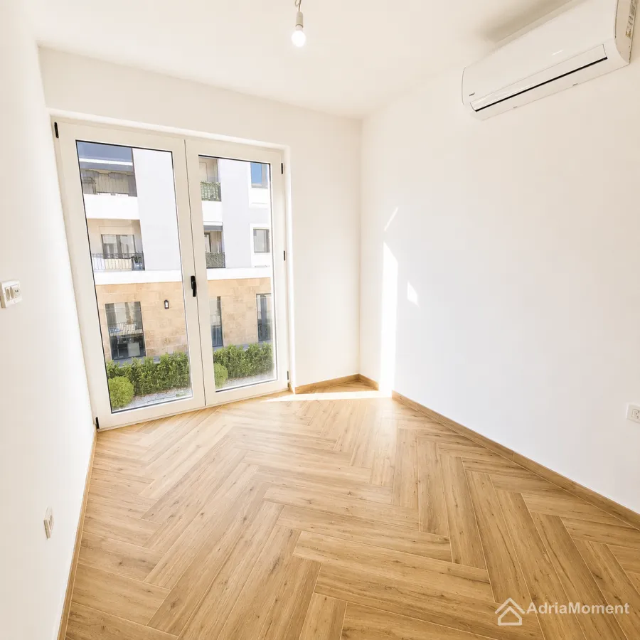 Prazna soba sa parketom u riblju kost, klima-uređajem i balkonskim vratima.