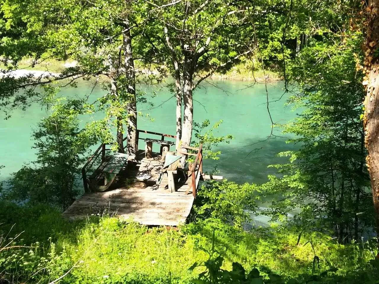 Drvena platforma sa klupama pored rijeke okružena bujnim zelenilom.