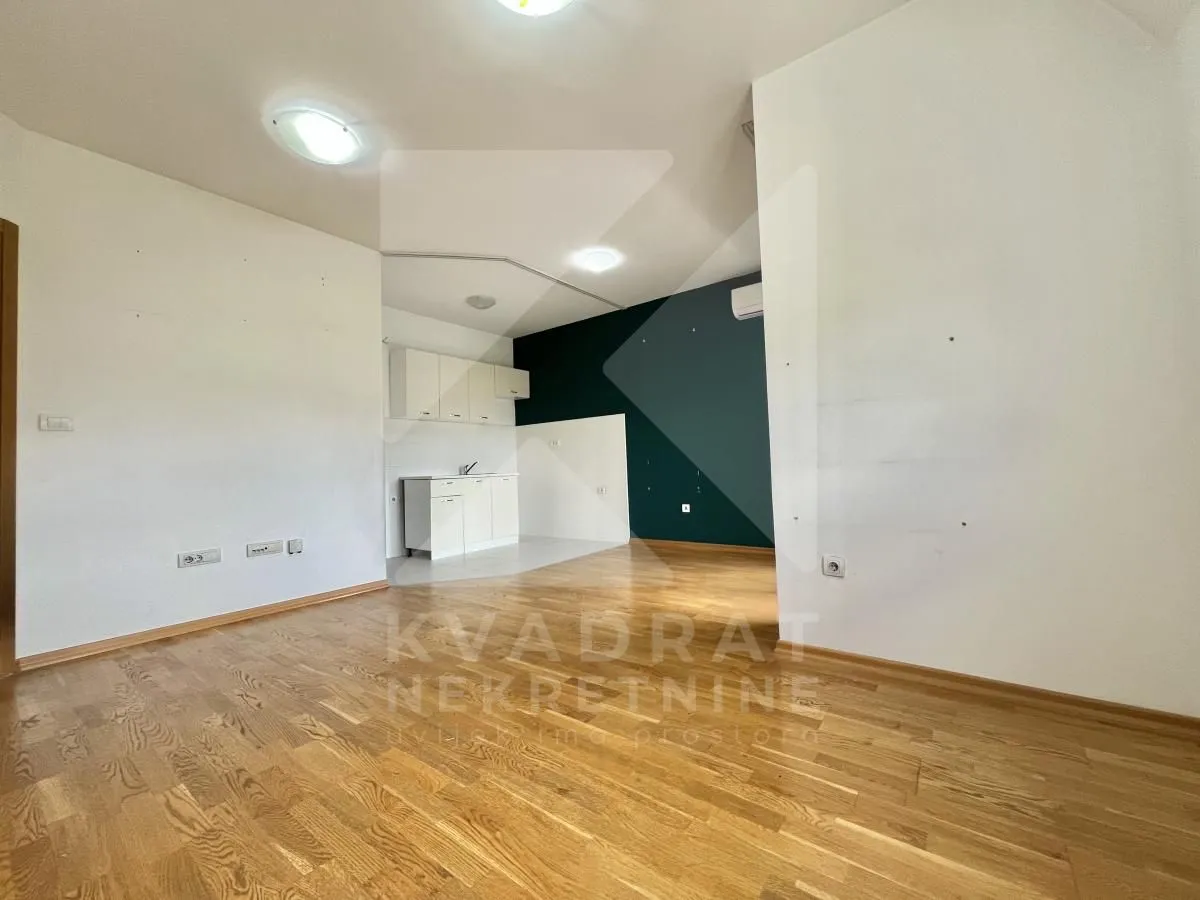 Empty living room with wooden flooring and a view of the kitchen in the background.