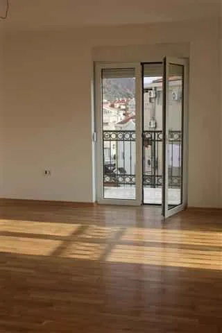 Empty room with parquet flooring and balcony doors overlooking neighboring buildings.