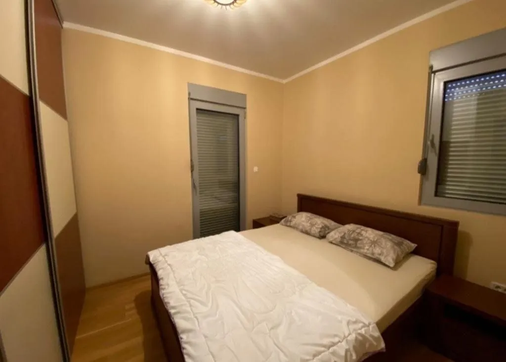 Namještena spavaća soba sa bračnim krevetom, ormarom i prozorima sa roletnama.