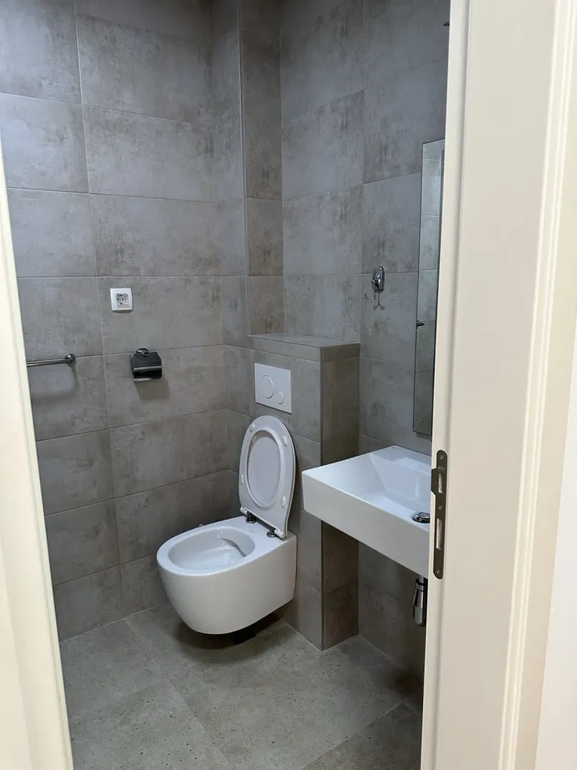 Modern half-bathroom featuring grey tiles, a toilet, and a small sink.