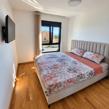 Svjetla spavaća soba sa bračnim krevetom, drvenim podom i televizorom na zidu. - image 4