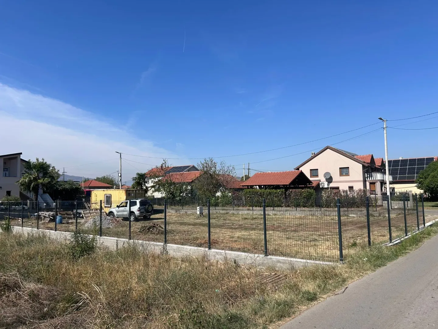 Pogled na ograđeni plac sa okolnim kućama i putem.