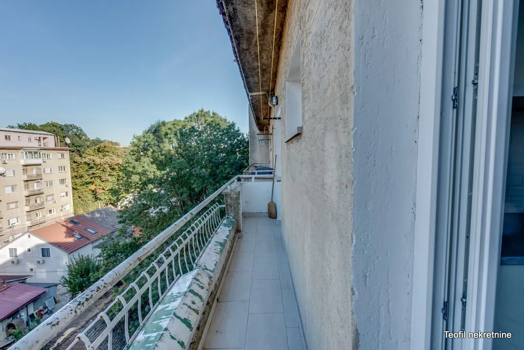 Dugačak popločani balkon sa metalnom ogradom i pogledom na naselje i zelenilo.