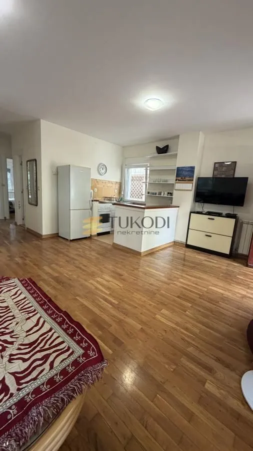 Living room with wooden flooring and a view of the kitchen in the background.