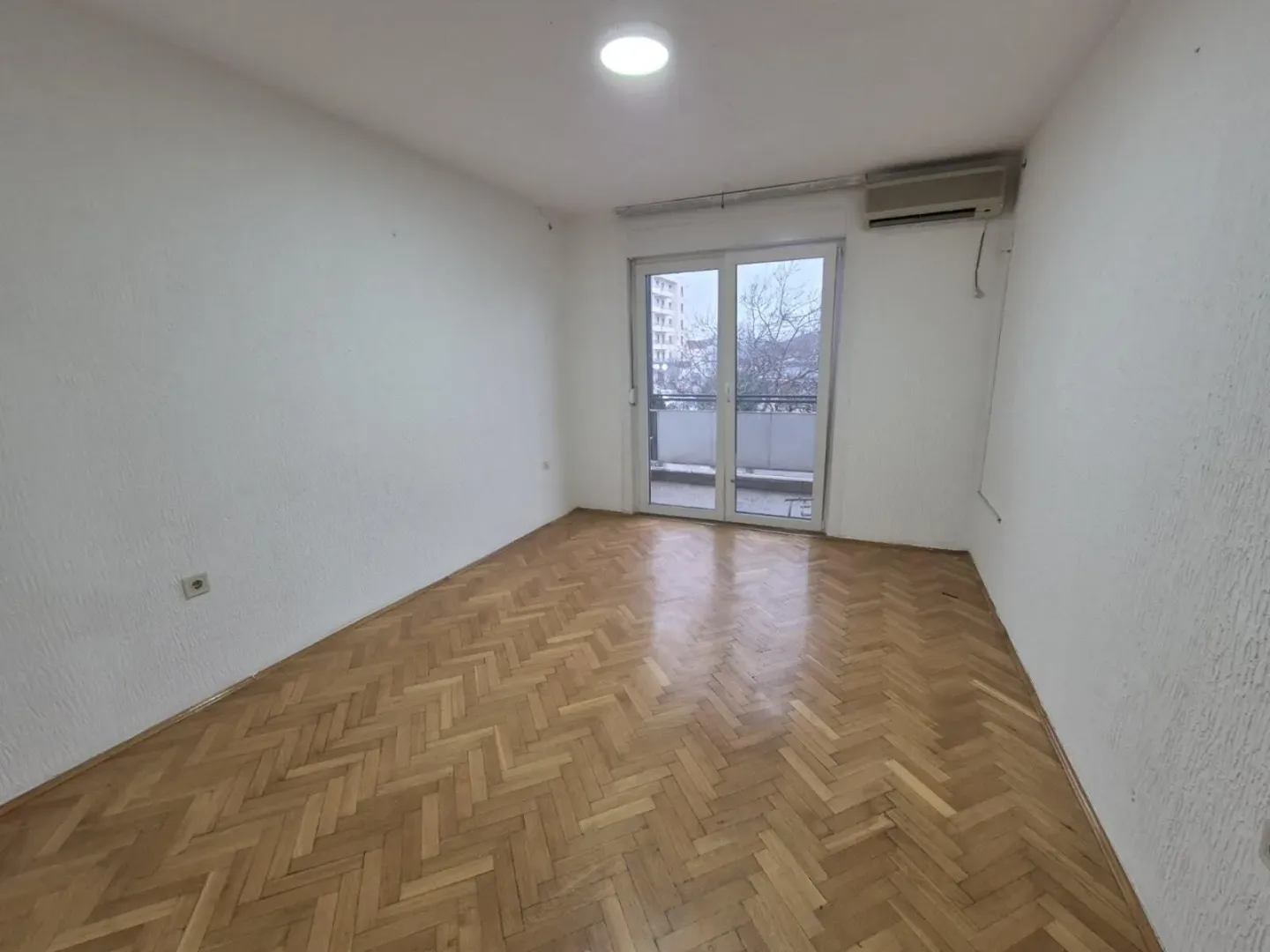 An empty, bright room with parquet flooring and balcony access.